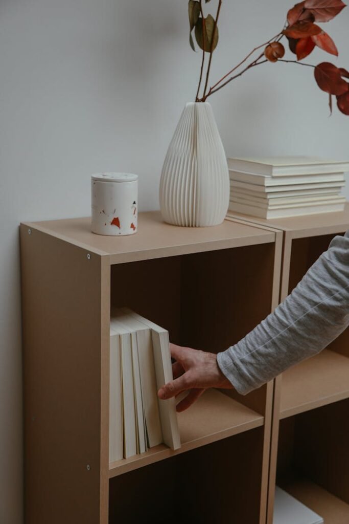 Une main tend la main vers un livre sur une étagère minimaliste, à côté d’un décor et de livres empilés.